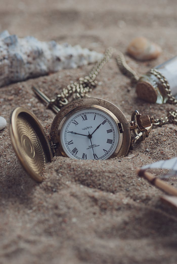 About A classic gold pocket watch partially buried in sand, exuding timeless elegance and a sense of nostalgia.